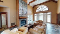 This impressive single-floor property in St. Andrews blends rustic charm with refined comfort through a thoughtfully designed open layout. The grand main living area immediately commands attention with its soaring vaulted ceiling accented by exposed wood beams and a dramatic floor-to-ceiling stone fireplace framed in rich wood trim. A large arched transom window floods the space with natural light, while French doors provide direct access to the outdoors, enhancing the airy, open atmosphere. Slate tile flooring and plush leather seating lend the room a warm, inviting character.

A second, more intimate lounge area transitions seamlessly from the main space through a wide wood-trimmed archway. This room features carpeted flooring, neutral toned walls, recessed lighting, and a ceiling fan, creating a relaxed media and gathering space complete with a flat-screen television and additional seating. Anchoring one corner is a substantial stacked-stone fireplace with a wood mantel, adding a cozy focal point to the room. A casual bar-height seating area nearby makes this space well-suited for entertaining.

Throughout the property, consistent use of natural wood accents, warm earth tones, and quality finishes creates a cohesive lodge-inspired aesthetic that is both functional and welcoming.