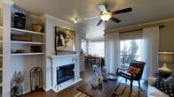 This single-story Flower Mound property showcases an open-concept layout that seamlessly connects the living and dining areas, creating a cohesive and functional living environment. The living room is anchored by a classic white-mantled fireplace, adding both warmth and a traditional focal point to the space. Crown molding trims the ceilings throughout, lending an elegant finishing touch to the interior. Dark hardwood flooring runs continuously through the main living areas, providing a rich contrast against the neutral gray walls and bright white trim. A ceiling fan with integrated lighting keeps the space comfortable and well-lit. Built-in shelving flanks the fireplace wall, offering display and storage opportunities. The dining area sits just beyond the living room, illuminated by a pendant light fixture and framed by large windows dressed with blinds and sheer white curtains, allowing for ample natural light. A sliding glass door opens to an outdoor balcony or patio, where views of mature trees enhance the sense of privacy and connection to the outdoors. The overall ambiance of the home is warm and inviting, blending classic architectural details with a comfortable, livable aesthetic.