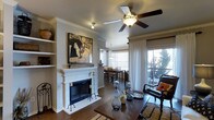 This single-story Flower Mound property showcases an open-concept layout that seamlessly connects the living and dining areas, creating a cohesive and functional living environment. The living room is anchored by a classic white-mantled fireplace, adding both warmth and a traditional focal point to the space. Crown molding trims the ceilings throughout, lending an elegant finishing touch to the interior. Dark hardwood flooring runs continuously through the main living areas, providing a rich contrast against the neutral gray walls and bright white trim. A ceiling fan with integrated lighting keeps the space comfortable and well-lit. Built-in shelving flanks the fireplace wall, offering display and storage opportunities. The dining area sits just beyond the living room, illuminated by a pendant light fixture and framed by large windows dressed with blinds and sheer white curtains, allowing for ample natural light. A sliding glass door opens to an outdoor balcony or patio, where views of mature trees enhance the sense of privacy and connection to the outdoors. The overall ambiance of the home is warm and inviting, blending classic architectural details with a comfortable, livable aesthetic.
