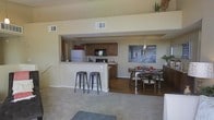 This single-story space is designed with an open-concept layout that seamlessly connects the kitchen, dining area, and living space. The kitchen is outfitted with medium-toned wood cabinetry, a built-in microwave, a full-size refrigerator, and light-colored countertops. A raised breakfast bar with two metal barstools extends from the kitchen, creating a casual seating option that also serves as a natural divider between the kitchen and the adjacent areas. The dining area sits just beyond the kitchen on hardwood-style flooring, accommodating a rectangular table with seating for several guests. A pendant light fixture hangs above the dining table, adding a focused lighting element, while a large monochrome wall art piece anchors the space visually. The living area features plush carpeting, contributing to a warm and comfortable atmosphere. Neutral, light-toned walls throughout enhance the sense of openness, and multiple ceiling vents suggest a central HVAC system. The overall layout is practical and functional, with clear zones for cooking, dining, and relaxing that flow naturally into one another.