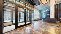 Located at 329 Alfred Ave in Teaneck, NJ, this expansive single-floor lobby space delivers a sophisticated, modern architectural experience designed to impress. The entrance features a grand glass-and-steel vestibule framed in warm wood accents, opening onto a stunning geometric marble-tile floor with dramatic veining in rich earth tones. Floor-to-ceiling windows and sheer drapery flood the interior with natural light, offering views of the surrounding wooded landscape.

The space unfolds across split levels connected by broad marble steps and glass-railed walkways. A prominent blue stone reception desk serves as a central focal point, accompanied by pendant globe lighting and exposed wood-beam ceiling details. A comfortable lounge area with curated seating provides an inviting gathering space nearby.

A standout biophilic installation features a lush moss wall and an interior garden bed with tropical foliage, bringing natural elements deep into the interior. Curved LED lighting traces the flowing ceiling lines throughout, enhancing the sense of movement and depth. Additional amenities include a billiards area with textured wall finishes and bar-height seating. Recessed and linear lighting accents architectural details throughout, creating a polished, upscale ambiance suited for a premier residential or commercial facility.