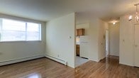 This single-floor space offers a straightforward and functional layout designed for comfortable everyday living. The main living area is finished with warm hardwood flooring that extends throughout, giving the space a cohesive and inviting feel. Neutral, light-toned walls keep the interior bright and airy, complemented by a window that allows natural light to fill the room. A baseboard heating system runs along the perimeter of the living area, ensuring year-round comfort. The kitchen is partially separated from the living space by a half-wall partition, providing a sense of definition while maintaining an open connection between the two areas. It features warm wood cabinetry, a white refrigerator, and a range, offering all the essentials for a fully functional cooking space. The kitchen is finished with lighter flooring, creating a subtle distinction from the living area. A hallway visible from the main space suggests additional rooms beyond, contributing to the overall flow and accessibility of the layout. The overall ambiance is clean, understated, and practical, making it a well-suited space for those seeking a no-frills, move-in-ready environment.