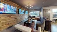 This single-floor communal space is designed with an urban-industrial aesthetic, combining raw materials with warm, eclectic furnishings to create a versatile social environment. Polished concrete floors extend throughout, providing a cohesive foundation beneath distinct activity zones.

The lounge area anchors one side of the space, featuring a striking reclaimed wood plank accent wall adorned with dual flat-screen televisions. Seating here includes a plaid upholstered sofa, wingback chairs, and a tufted bench arranged around a central coffee table, all bathed in natural light from large windows overlooking an outdoor courtyard. An architectural chandelier adds a mid-century modern touch overhead.

Adjacent to the lounge, a generously sized dining area accommodates a long wooden table surrounded by leather and metal chairs, illuminated by a statement cluster pendant light. Plaid drapes frame the windows, softening the industrial palette. A shuffleboard table and foosball table provide additional recreational options within the open layout.

The entry zone features warm wood-paneled walls with pendant lighting, a small bar or service counter, and open sightlines across the entire space. Direct access to an exterior courtyard is provided through glass-paneled doors, connecting the interior to outdoor areas and enhancing the flow of natural light throughout.