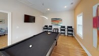 This game room and lounge area is thoughtfully designed to offer both active entertainment and relaxed social gathering in a single, well-appointed space. The layout flows naturally between two distinct zones, each with its own character and purpose.

The game room section centers around a full-size pool table with a black felt surface, complemented by a sleek arc pendant light overhead and a wall-mounted flat-screen TV. High bar-style seating lines the wall, providing spectator spots alongside a dedicated cue rack. Vibrant, colorful artwork adorns the walls, and warm-toned accent walls add energy to the room. Multiple windows along the exterior wall invite natural light and offer views of the greenery outside, while light hardwood flooring ties the space together seamlessly.

The lounge area transitions effortlessly from the game room, featuring impressively soaring ceilings that lend an airy, open feel to the space. Comfortable blue upholstered armchairs are arranged for easy conversation, set against neutral walls accented with framed artwork. A large wall-mounted TV serves as a focal point, and glass-paned doors connect to an adjacent dining or gathering area, enhancing the sense of openness. The consistent hardwood flooring throughout unifies both zones, creating a cohesive and inviting environment perfect for socializing and recreation.