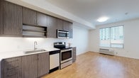 This well-designed single-floor unit offers a clean, contemporary layout that maximizes functionality and comfort. The open-concept living and kitchen area forms the heart of the space, where warm hardwood flooring flows seamlessly underfoot, creating a bright and inviting atmosphere. A large window allows natural light to fill the living area, offering views of the surrounding urban environment and enhancing the overall sense of openness.

The kitchen is outfitted with rich, dark wood-grain cabinetry that extends to the ceiling, providing generous storage capacity. Stainless steel appliances — including an electric range, over-the-range microwave, and dishwasher — complement the light countertops, striking a modern contrast against the cabinetry. Open shelving adds both practicality and visual interest to the kitchen design.

The full bathroom features a combined tub and shower, a toilet, and a vanity with a single basin sink and matching wood-grain cabinetry, all finished in a clean, neutral palette. A large mirror above the vanity adds brightness to the room.

Additional highlights include a spacious walk-in closet equipped with wire shelving and hanging rods for versatile storage, as well as a dedicated laundry area with an in-unit washer and dryer. The overall design is cohesive, modern, and thoughtfully appointed throughout.