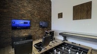 This single-floor entertainment space is purpose-built for recreation and social play, offering a compelling mix of gaming options and bold interior design. The room's most striking feature is a dramatic floor-to-ceiling stacked stone accent wall in rich, earthy tones of brown and charcoal, which provides a strong textural contrast against the clean white walls on the opposing side. A wall-mounted flat-screen television is positioned prominently on the stone wall, accompanied by an arcade game cabinet that adds a nostalgic, classic gaming touch. Decorative wall-mounted wooden art pieces add warmth and character to the white walls. The space is grounded by light hardwood flooring that balances the room's darker elements. A foosball table takes center stage in the foreground, while an air hockey table is positioned nearby, making the layout ideal for group entertainment. A comfortable black leather gaming chair completes the setup, offering a dedicated seating area in front of the screens. The overall ambiance is energetic yet refined, blending rustic stone textures with modern entertainment technology to create a well-equipped, inviting game room.