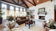 This single-story clubhouse showcases a refined Hill Country aesthetic, blending natural materials with thoughtful design throughout its welcoming interior. The grand entryway greets visitors with large-format slate tile flooring in earthy tones, accented by rich wood trim and a striking natural stone column that anchors the space. Soaring ceilings with warm, stained wood paneling and exposed dark beams carry throughout the main areas, lending the space an open, airy quality while maintaining a cozy, lodge-like atmosphere.

The main lounge area is a focal point of the space, anchored by a floor-to-ceiling stacked stone fireplace with a wood mantel. Comfortable leather seating and a patterned area rug create an inviting gathering environment. Tall windows dressed in layered curtains allow generous natural light to filter in while offering views of the surrounding exterior.

Adjacent areas are accessible through elegant wood-and-glass French doors, revealing what appears to be a business center and additional lounge space, each maintaining consistent design elements including recessed lighting, wood-beamed ceilings, and warm wall sconces. The overall ambiance is sophisticated yet comfortable, balancing rustic charm with polished finishes.