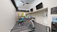 Located at 3500 Windmeadows Blvd in Gainesville, Florida, this fitness center is a well-equipped, single-floor exercise facility designed to support a variety of workout routines. The space features soaring ceilings with clerestory windows that allow natural light to pour in from above, creating an open and energizing atmosphere. A ceiling fan helps maintain comfortable airflow throughout the room, complementing the HVAC vents positioned along the upper walls.

The main workout area is finished with durable rubber flooring and lined with full-length mirrors along one wall, enhancing the sense of space while allowing users to monitor their form during exercise. The room accommodates a treadmill and an adjustable workout bench as primary cardio and strength-training stations. A dedicated rack organizes a full set of dumbbells, while a multi-purpose storage unit holds an assortment of medicine balls, stability balls in varying sizes, resistance bands, and foam rollers — offering a comprehensive range of functional fitness options.

Two wall-mounted flat-screen televisions provide entertainment during workouts. A water cooler station and waste receptacles are conveniently positioned near the entrance. Posted exercise room rules ensure a safe and respectful environment for all users. The entry area features terracotta tile flooring and a glass-paned French door, providing a welcoming transition into the facility.