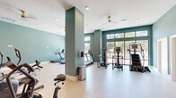 This modern fitness center offers a generously sized, single-floor layout designed to accommodate a comprehensive range of workout equipment and training styles. The space is defined by its contemporary aesthetic, featuring soothing teal and sage green accent walls paired with light wood-style flooring that creates a clean, energizing atmosphere throughout.

The main workout area is anchored by a row of treadmills positioned directly in front of expansive floor-to-ceiling windows, providing outdoor views and abundant natural light during cardio sessions. Additional cardio equipment includes elliptical trainers, stationary cycling bikes, and a rowing machine, offering users a full spectrum of aerobic options. Ceiling fans and recessed lighting ensure comfort and consistent illumination across the space.

A standout architectural feature is the glass-walled enclosure framed in sleek black steel, which houses a dedicated strength training and weight area. This transparent partition maintains an open, airy feel while visually and functionally separating the zones. A wall-mounted TV screen is also present for entertainment during workouts.

Structural columns are integrated into the design without disrupting usability, and the high ceilings contribute to a spacious, unenclosed feel. The overall layout is thoughtfully organized to allow users to move freely between equipment, making this fitness center both functional and visually inviting.