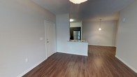 This single-floor residence showcases a clean, modern design characterized by neutral gray walls and rich hardwood-style flooring that flows continuously throughout the living areas, creating a cohesive and welcoming atmosphere. The entryway opens directly into the main living space, offering a sense of openness and ease of movement. A partial wall with a pass-through counter connects the kitchen to the living area, maintaining an open-concept feel while subtly defining each zone. The kitchen is equipped with stainless steel refrigerator and features dark cabinetry that provides a striking contrast against the lighter walls and countertops. Overhead lighting in the kitchen is complemented by a stylish pendant light in the living area, adding a contemporary decorative element. White trim and baseboards frame the space neatly, contributing to a crisp, well-finished appearance. The layout is practical and functional, with the entry door positioned conveniently along the side wall. Overall, the unit presents a straightforward, modern aesthetic with thoughtful design choices that balance style and everyday usability.