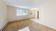 Located at 100 Almeida Terr in Portsmouth, Rhode Island, this single-floor unit offers a straightforward, functional layout with a clean, neutral aesthetic throughout. The galley-style kitchen is efficiently designed with white cabinetry, ample upper and lower storage, white appliances including a refrigerator, electric range, and built-in microwave, along with a stainless steel sink and marble-look tile flooring. The warm, cream-toned walls carry through the living areas, where plush carpeting and baseboard heating create a comfortable environment. Multiple living and bedroom spaces feature generous windows that invite natural light and offer views of the surrounding greenery and outdoor landscape. One bedroom includes a dedicated closet and large windows overlooking a natural rock feature, adding a distinctive exterior element. A chandelier-lit dining or entry area connects the main living space, providing an open transition point. The full bathroom is neatly appointed with white subway tile surrounding a soaking tub, a vanity with storage, a mirrored medicine cabinet, and matching marble-look tile flooring. A wall-mounted air conditioning unit is present in one of the rooms, supplementing the baseboard heating system. The overall ambiance is bright, clean, and move-in ready.