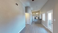 This single-story, 2-bedroom, 2-bathroom home showcases an open-concept layout that seamlessly connects the living area to the kitchen. The space immediately impresses with its vaulted ceilings, which lend an airy, expansive feel to the interior. Light gray walls and light-toned wood-look flooring run throughout, creating a clean, cohesive aesthetic.

The living area is generously proportioned and benefits from natural light streaming in through a side window, complemented by a ceiling fan overhead. A rounded archway detail adds architectural character, providing a smooth visual transition between spaces.

The kitchen is positioned at the rear of the open-plan area and features light wood cabinetry, dark countertops, and white appliances, including a refrigerator, microwave, and range. Pendant lighting hangs above, illuminating the space with a warm glow. A kitchen bar or peninsula offers additional counter space and casual seating potential.

A louvered bi-fold door, likely concealing a closet or utility space, is visible along the hallway leading toward the bedrooms and bathrooms. The overall ambiance is bright, clean, and functional, with thoughtful architectural details that enhance the sense of openness.