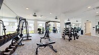 This modern fitness center in Redlands, CA is thoughtfully designed to accommodate a full range of workout disciplines across a generous single-floor layout. The main exercise area features neutral-toned carpet flooring, white structural columns, and an abundance of natural light streaming through large windows that offer views of the surrounding outdoor areas. Recessed lighting and multiple ceiling fans ensure a bright, comfortable environment throughout.

The strength training zone is well-appointed with a multi-station cable machine, a comprehensive dumbbell rack, weight benches, and additional resistance equipment. The cardio area includes treadmills, elliptical trainers, stair climbers, and stationary bikes, all positioned near the expansive windows. Wall-mounted flat-screen televisions are conveniently placed throughout the space to keep users engaged during workouts.

A dedicated studio room sets itself apart with warm hardwood flooring, a bold geometric-patterned accent wall, floor-to-ceiling mirrors, and its own flat-screen TV — ideal for yoga, stretching, spin classes, or group fitness sessions. The studio connects seamlessly to the main floor via a frosted-glass interior door. The overall design strikes a balance between clean, contemporary aesthetics and functional fitness infrastructure, making this facility a comprehensive destination for health and wellness.