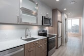 This contemporary single-floor unit is thoughtfully designed with a clean, modern aesthetic throughout. The galley-style kitchen is efficiently laid out and outfitted with stainless steel appliances, including an electric range, over-the-range microwave, dishwasher, and refrigerator. Two-tone cabinetry — combining frosted glass upper cabinets with wood-toned lower units — pairs with white quartz countertops and a stainless steel undermount sink, creating a polished, functional workspace. Light wood-tone flooring runs seamlessly through the unit, adding warmth and visual continuity. The bathroom features a well-lit LED backlit vanity mirror, wood-toned upper cabinetry, a white quartz vanity top, and a full tub-shower combination enclosed with a glass door and tiled surround. A stacked in-unit washer and dryer provides added convenience. The bedroom is generously lit by natural light and includes a sliding closet and direct access to the bathroom. The living area is highlighted by a large floor-to-ceiling window framing open city views, allowing abundant natural light to fill the space. Neutral wall tones and recessed lighting throughout contribute to the unit's bright, contemporary ambiance.