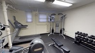 This fitness center offers a practical and functional workout space outfitted with a comprehensive selection of exercise equipment to support a variety of training routines. The space is illuminated by overhead fluorescent lighting and features neutral-toned walls with rubber floor matting laid throughout the exercise areas to provide stability and protection.

The cardio section includes a Precor elliptical trainer, a recumbent bike, and a treadmill, catering to a range of cardiovascular workout preferences. A multi-function cable crossover machine serves as the centerpiece of the strength training area, enabling a wide variety of resistance exercises. Complementing this is a dumbbell rack stocked with a selection of free weights, a flat utility bench, and a medicine ball rack. Exercise balls in multiple sizes add flexibility for core and balance training.

A small set of windows along one wall allows for modest natural light, while exposed utility piping runs along the ceiling, giving the space a straightforward, no-frills aesthetic. A wall-mounted electrical panel and keypad entry system are also present. The overall layout is efficient and purposeful, making effective use of the available floor space to accommodate multiple users and exercise modalities simultaneously.