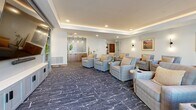 Located at 6504 N 67th Ave in Glendale, Arizona, this well-appointed communal living room offers a welcoming and versatile gathering space. The room features a tray ceiling with recessed lighting and warm ambient cove lighting that creates a polished, refined atmosphere throughout.

The space is anchored by a large wall-mounted flat-screen television set within a built-in media console with ample cabinetry storage below, making it ideal for group viewing and entertainment. Generous seating is arranged thoughtfully across the room, including plush upholstered armchairs and sofas in neutral tones, accented with warm throw pillows and paired with small round side tables for added functionality.

A patterned blue-grey carpet with abstract white detailing covers the floor, adding texture and visual interest. Along one wall, a built-in wet bar with pendant lighting, a tile backsplash, and warm wood cabinetry provides a convenient refreshment station.

Large sliding glass doors and windows along one side of the room flood the interior with natural light while offering views of the outdoor greenery and surrounding landscape. A built-in bookcase with curated décor and framed Arizona-themed artwork contribute to the space's warm, community-centered character. Stylish wall sconces and thoughtful touches throughout complete this inviting, functional gathering room.