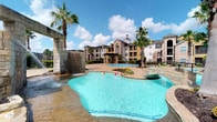This outdoor pool area delivers a resort-style experience within a residential community setting. The centerpiece is a generously sized freeform pool with striking waterfall features built into a stone-clad structure, sending cascading water into the crystal-clear turquoise basin below. The pool's organic shape adds a naturalistic feel, complemented by colorful landscaping and tropical palm trees strategically placed throughout the deck area.

The expansive concrete pool deck provides ample space for lounging and recreation, with a textured, slip-resistant finish. Stone accent columns and architectural details tie the outdoor environment to the surrounding buildings, which rise in the background with warm stucco and stone facades typical of a Southwestern-influenced design.

Adjacent to the pool is a clubhouse or amenity building featuring a covered entrance with prominent natural stone columns, crisp white board-and-batten siding, and large floor-to-ceiling windows framed by a grand arched transom. This covered corridor connects the interior facilities to the pool deck, offering a shaded transitional space. The floor-to-ceiling glazing allows abundant natural light to flood the interior while providing unobstructed views of the pool area. Together, these elements create a cohesive, welcoming outdoor amenity space designed for both relaxation and social activity.