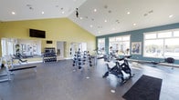The fitness center is a spacious, thoughtfully designed facility that combines functionality with an open, energizing atmosphere. The space is defined by its dramatic vaulted ceiling, which rises to a striking peak and is fitted with recessed lighting and a ceiling fan, ensuring the area remains well-lit and comfortable during workouts. The walls are painted in a two-tone palette of warm yellow and deep teal, creating a vibrant and motivating environment.

Natural light pours in through a row of large windows along one wall, offering views of the surrounding exterior and adding to the bright, airy feel of the space. The durable, dark rubberized flooring provides a safe and practical surface throughout.

Equipment is well-distributed across the floor, including a rack of free weights and dumbbells along one wall, a multi-function weight rack, spinning/cycling bikes in the foreground, and a lineup of cardio machines—including treadmills and ellipticals—positioned near the windows. A wall-mounted television is also present. An adjacent doorway suggests access to additional amenities nearby.