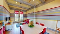 This cheerful children's playroom is designed with young minds in focus, offering a lively and stimulating environment across a single open floor. The most striking architectural feature is the bold horizontal striped wall treatment in yellow, gray, and red that wraps the entire room, creating an energetic and visually engaging atmosphere. Exposed ceiling beams add structural character overhead, while recessed lighting and a ceiling fan ensure the space remains comfortable and well-lit.

The room is divided into distinct activity zones. A central activity table surrounded by bright red chairs serves as the primary creative and learning hub, topped with a colorful toy centerpiece. Adjacent to it, a white open bookshelf stores educational toys, an abacus, and activity materials, keeping essentials organized and accessible.

A cozy lounge area features a bold red sofa accented with yellow throw pillows, positioned atop a playful blue handprint area rug — ideal for storytime or relaxed play. A media wall with mounted flat-screen televisions and a low storage console provides an entertainment zone, complemented by additional toy storage bins nearby.

Large windows dressed with red roller shades flood the space with natural light and offer views of lush outdoor surroundings including palm trees. Glass double doors provide direct access to an outdoor courtyard, seamlessly connecting indoor and outdoor play environments.