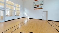 Located at 2525 Crescent View Dr in Charlotte, NC, this dedicated indoor basketball court is a standout recreational facility designed for serious play and active enjoyment. The court features a classic hardwood maple floor with regulation court markings, including a painted key, center circle, and boundary lines, providing an authentic playing surface. A wall-mounted, fold-down glass backboard with a standard rim and net anchors one end of the court, offering a professional-grade playing experience.

The space is defined by its impressively soaring ceilings, which provide more than ample vertical clearance for gameplay. Crisp white walls lend a clean, bright aesthetic throughout. Natural light floods the interior through multiple large, multi-pane windows positioned along the walls—including a dramatic floor-to-ceiling window bank that frames views of the surrounding neighborhood—creating a welcoming and energetic atmosphere.

Access to the court is provided through a set of double glass doors framed by interior transom windows, which also allow visibility into the adjacent space. Additional interior-facing windows further connect the court to neighboring rooms. A decorative wall plaque adds a personal touch to the space. The overall design is both functional and well-appointed, making this indoor court a truly exceptional feature of the property.