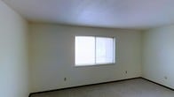This single-floor space presents a straightforward and functional interior. The room visible in the image features neutral-toned walls that provide a clean, understated backdrop, complemented by wall-to-wall carpeting that adds a degree of warmth underfoot. A centrally positioned window fitted with horizontal blinds allows natural light to filter into the room, brightening the space and offering a view of the surrounding outdoor environment. The textured ceiling adds a subtle visual detail to the otherwise minimalist interior. Electrical outlets are positioned along the lower portion of the walls, contributing to the room's practical utility. The overall design is modest and unadorned, making it a versatile space adaptable to a variety of uses. The light, neutral color palette throughout creates a cohesive and calm atmosphere.