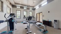 This fitness center offers a bright, open workout environment defined by soaring ceilings and an elevated clerestory window design that floods the space with natural light from multiple directions. The neutral-toned walls and speckled flooring create a clean, professional atmosphere throughout the single-level layout.

The cardio section is well-appointed with multiple treadmills equipped with individual monitor screens, an upright stationary bike, an elliptical trainer, a stair climber, and a rowing machine — providing a thorough range of aerobic training options. Teal exercise mats are strategically placed throughout the floor to designate workout zones.

The strength training area features a multi-functional cable machine suitable for a wide variety of resistance exercises, a dumbbell rack with a full set of free weights, and an adjustable weight bench. Full-length mirrors line one wall, enhancing the sense of space and allowing users to monitor their form during workouts.

A ceiling fan provides additional air circulation, and a water dispenser is conveniently located near the exit. Two sets of French doors open to an outdoor area, offering easy access and additional ventilation. The overall design balances functionality with an inviting, well-lit ambiance suited for a comprehensive fitness routine.