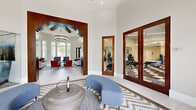 Located in Tampa, FL, this beautifully designed single-floor clubhouse facility offers a refined and welcoming atmosphere across a generously sized layout. The grand entrance foyer greets visitors with soaring ceilings, a dramatic arched doorway framed in rich dark wood, and a striking chevron-patterned tile floor. Curved blue benches flank a sculptural metallic centerpiece table, setting an elegant tone from the moment of arrival. Floor-to-ceiling arched windows allow abundant natural light to pour in, offering views of the lush, landscaped grounds outside. The spacious living room is a centerpiece of the space, anchored by a large tiered tray ceiling with exposed dark wood beam detailing and an impressive double-ring chandelier. Comfortable seating arrangements in neutral and earth tones invite relaxed gathering, while a marble-clad accent wall houses a sleek linear fireplace and wall-mounted display. An adjacent dining and lounge area features pendant lighting and a wet bar. Glass barn doors and wood-framed French doors lead to a well-equipped fitness center, visible from the main hall and finished with bold checkered flooring. Throughout, the design blends modern sophistication with warm architectural accents, creating a polished and inviting communal space.