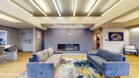 Located at 1685 Athena Dr in Kent, Ohio, this single-floor community space offers a well-appointed common area designed for gathering and relaxation. The layout encompasses a living room, dining area, and kitchen, all finished with light hardwood flooring that flows seamlessly throughout.

The living room serves as the centerpiece of the space, anchored by a sleek, wide linear fireplace set into a bold blue-gray textured accent wall. Plush blue velvet sofas and a vibrant abstract area rug in blue, teal, and gold add a contemporary, spirited character to the room. A coffered ceiling with integrated linear lighting fixtures adds architectural depth overhead. Large glass entry doors at one end of the living area invite abundant natural light and offer views of the outdoor greenery beyond.

A wide hallway connects the main lounge to additional areas, decorated with Kent State University branding that reinforces the community's collegiate identity. The kitchen features crisp white cabinetry, a bright quartz countertop, and a spacious island with bar seating for multiple guests. A large picture window above the counter frames views of the surrounding campus landscape. An adjacent kitchenette with additional cabinetry and a utility sink provides extra functionality. Access to a fitness center is also available directly from this space.