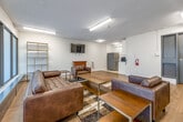 This single-floor communal space is thoughtfully designed to serve as a comfortable and functional gathering area. The main lounge is bright and open, featuring light-toned hardwood flooring throughout and a textured ceiling with flush-mounted fluorescent lighting that ensures consistent illumination across the room. A large bank of frosted floor-to-ceiling windows lines one wall, allowing diffused natural light to fill the space while maintaining privacy. A wall-mounted split air conditioning unit provides climate control.

The lounge is furnished with a set of rich brown leather sofas and armchairs arranged around a gold-framed coffee table, all anchored by a patterned area rug in earth tones. A wall-mounted flat-screen TV is positioned above a compact electric fireplace, creating a welcoming focal point. An open shelving unit adds a decorative touch, while abstract wall art near the entrance contributes a contemporary aesthetic.

Adjacent to the main lounge is a compact kitchenette, finished with ceramic tile flooring, white cabinetry, a stainless steel sink, a microwave, and a full-size refrigerator — providing convenient amenities for occupants. A secured entry door with safety signage ensures controlled access to the space, making this a practical and inviting communal environment.