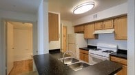 This single-floor space centers around a practical and well-appointed kitchen that serves as its defining feature. The kitchen is outfitted with dark speckled stone countertops that provide ample workspace, complemented by warm light wood cabinetry offering generous storage above and below. A double stainless steel sink sits prominently at the kitchen peninsula, making it a functional hub for meal preparation. White appliances, including a gas range and a full-size refrigerator, are neatly integrated into the layout alongside a range hood. Overhead, a flush-mount circular ceiling light provides bright, even illumination throughout the space. The kitchen connects to adjacent areas, including what appears to be a bathroom visible to one side and additional rooms accessible through doorways, suggesting a compact but complete floor plan. Neutral-toned walls and standard flooring run throughout, giving the space a clean, understated appearance. The layout is straightforward and efficient, making good use of the available space while keeping all essential living elements within easy reach on a single level.