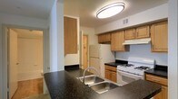 This single-floor space centers around a practical and well-appointed kitchen that serves as its defining feature. The kitchen is outfitted with dark speckled stone countertops that provide ample workspace, complemented by warm light wood cabinetry offering generous storage above and below. A double stainless steel sink sits prominently at the kitchen peninsula, making it a functional hub for meal preparation. White appliances, including a gas range and a full-size refrigerator, are neatly integrated into the layout alongside a range hood. Overhead, a flush-mount circular ceiling light provides bright, even illumination throughout the space. The kitchen connects to adjacent areas, including what appears to be a bathroom visible to one side and additional rooms accessible through doorways, suggesting a compact but complete floor plan. Neutral-toned walls and standard flooring run throughout, giving the space a clean, understated appearance. The layout is straightforward and efficient, making good use of the available space while keeping all essential living elements within easy reach on a single level.