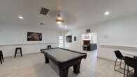 This single-floor community clubhouse is designed to offer a versatile and inviting gathering space across several distinct areas. The game room serves as a central highlight, featuring a full-size pool table set against crisp white walls adorned with billiards-themed artwork. Wall-mounted cue racks, bar-height stools, and a decorative ceiling fan complete the recreational atmosphere, all set atop light wood-look tile flooring that flows consistently throughout the space.

Adjacent to the game room, the lounge area provides a comfortable retreat with two substantial leather sofas arranged around a set of round nesting coffee tables. A wood-mantled fireplace anchors the room, flanked by built-in shelving units, while roller shades on large windows allow for controlled natural light. A ceiling fan and recessed lighting ensure the area remains bright and well-ventilated.

A dedicated dining area offers multiple round tables with modern seating, bathed in natural light from large windows fitted with sheer roller shades. A decorative indoor plant adds a touch of greenery to the clean, minimal aesthetic.

The kitchen is fully functional, featuring warm wood cabinetry, marble-look countertops, a sink, and a full suite of black appliances including a stove, microwave, and refrigerator — well-suited for catering or community events.