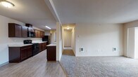 Located at 1090 North Cecil Road in Post Falls, Idaho, this single-floor residence offers a thoughtfully designed layout with a seamless flow between living spaces. The open-concept kitchen and living area serves as the heart of the unit, where wood-look hard surface flooring transitions into plush carpeting in the living room. The kitchen is outfitted with rich dark-stained cabinetry, light marble-style countertops, and black appliances including a range, microwave, and refrigerator, with generous counter space lining both sides of the galley-style layout. A peninsula provides additional workspace and a natural divide between the kitchen and living area. The living room features a wall-mounted air conditioning unit and direct access to a private balcony with exterior views. A hallway leads to the bedrooms, each carpeted and filled with natural light from sizeable windows. One bedroom features a ceiling fan and direct access to an en suite bathroom. The bathrooms are finished consistently with dark wood vanities, light stone-look countertops, and wood-look tile flooring. The full bathroom includes a tub-shower combination, while the secondary bathroom offers a single-sink vanity. Neutral wall tones and clean lines give the entire space a bright, move-in-ready feel.