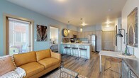 This single-floor unit offers a thoughtfully designed open-concept layout where the living area and kitchen flow seamlessly together. The living room is bathed in natural light through large windows fitted with horizontal blinds, offering views of the surrounding neighborhood. Wood-look flooring runs throughout the main living spaces, complementing the soft blue-gray accent walls that lend the unit a calm, contemporary feel.

The kitchen is well-equipped with light wood cabinetry, stainless steel appliances—including a refrigerator, range, and microwave—and a white countertop peninsula with bar seating for casual dining. Pendant lighting above the peninsula adds a refined touch.

The primary bedroom features carpeted flooring, multiple windows for natural light, and ample space for furnishings. A secondary bedroom offers similar comfort with carpeted floors, a window, and direct access to a bathroom. The full bathroom includes a light wood vanity with white countertops, a tub-shower combination, and wood-look flooring.

A dedicated laundry room houses a full-size washer and dryer alongside built-in shelving for storage. Throughout the unit, the cohesive use of light wood tones, neutral walls, and modern fixtures creates a clean, functional living environment.