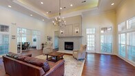 Located in Germantown, TN, this single-story leasing center presents a polished, upscale environment designed to welcome visitors and showcase community living. The main lounge is the architectural centerpiece, boasting soaring tray ceilings adorned with detailed crown molding and illuminated by a striking multi-arm chandelier. Rich hardwood flooring extends throughout, complementing the warm, neutral color palette. A floor-to-ceiling fireplace surround, finished in soft gray paneling with built-in cabinetry above, anchors the room and serves as a dramatic focal point. Comfortable leather seating and patterned accent chairs are arranged around a wood coffee table atop a layered area rug, creating an inviting, residential atmosphere.

Large French doors and multi-pane windows flood the lounge with natural light and offer views of the surrounding greenery. Adjacent to the main seating area, a dedicated leasing office features a dark wood desk, open shelving for displays, and a small beverage station — providing a functional yet refined workspace. A glass-enclosed fitness center is also accessible from the common area, equipped with cardio machines and exercise equipment, adding a practical amenity within the same footprint. Overall, the space balances elegance with functionality.