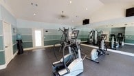 Located at 51 Northtown Dr in Jackson, Mississippi, this single-floor fitness center offers a well-equipped and thoughtfully designed workout space. The room features vaulted ceilings that lend an open, airy feel, complemented by recessed lighting and a ceiling fan for comfort during workouts. The walls are painted in layered tones of teal and white, creating a calm, motivating atmosphere throughout the space.

A full wall of floor-to-ceiling mirrors runs along one side of the room, visually expanding the space and allowing users to monitor their form during exercise. Large French doors with glass panels line the opposite wall, inviting abundant natural light and offering views of the surrounding greenery outside.

The fitness center is outfitted with a diverse selection of equipment, including elliptical trainers, a treadmill, a stationary bike, and a multi-function cable strength machine, accommodating both cardio and resistance training needs. Protective rubber flooring with interlocking mat sections covers the workout area, providing safety and durability underfoot.

Additional amenities include a wall-mounted flat-screen television for entertainment during workouts and a water cooler station near the entrance. The overall design strikes a balance between functionality and comfort, making this fitness center a practical and inviting facility for residents of the Northtown Dr community.