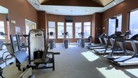 This single-floor fitness center offers a thoughtfully designed workout environment that balances functionality with comfort. The space is defined by its warm, deep brown accent walls and a striking vaulted ceiling with wood detailing, lending the room an upscale, welcoming atmosphere. Large windows line the far wall, flooding the interior with abundant natural light and offering views of the surrounding greenery, creating an energizing backdrop for exercise. The cardio area is well-appointed with multiple elliptical trainers and treadmills arranged to maximize space and usability. Strength training equipment, including a multi-function weight machine with an adjustable bench, is positioned toward the front of the room, catering to a range of fitness goals. Recessed lighting supplements the natural light, ensuring the space remains bright and functional at all times. A wall-mounted flat-screen television is centrally positioned, providing entertainment during workouts. The flooring features durable, neutral-toned carpet suited for a fitness environment. Overall, the space presents a clean, organized, and fully equipped facility designed to support a comprehensive fitness routine in a comfortable and visually appealing setting.