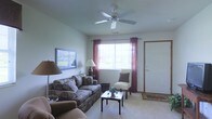 This single-floor space centers around a welcoming living room that combines comfort and functionality. The room is carpeted throughout, providing a warm underfoot feel, and features neutral-toned walls that create a clean, understated backdrop. A ceiling fan is mounted overhead, offering both air circulation and subtle illumination to the space.

Natural light flows in through a large central window dressed with rich burgundy curtains, brightening the room and providing a view of the outdoors. The living area is furnished with a patterned sofa and a coordinating armchair, arranged around a rectangular coffee table that anchors the seating arrangement. An additional accent chair near the window adds versatility to the layout.

Supplementary lighting is provided by a classic floor lamp and a matching table lamp, ensuring the space remains well-lit during evening hours. A framed artwork piece on the wall adds a personal touch to the décor. The front entry door, positioned to one side of the room, is flanked by a small welcome mat, seamlessly integrating the entryway into the overall living space. The overall ambiance is relaxed and livable, suited to everyday comfort.