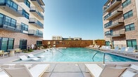 This outdoor amenity space is a thoughtfully designed resort-style pool courtyard nestled within a contemporary multi-story residential complex. The centerpiece is a large, shimmering swimming pool with crystal-clear blue water, flanked on multiple sides by the building's modern facade — a striking combination of smooth white stucco and warm brick detailing. Private balconies from the surrounding upper floors overlook the pool area, creating an immersive courtyard feel.

The pool deck is generously proportioned and finished with light-toned concrete, offering ample space for relaxation. A standout feature is the row of in-water loungers positioned along a shallow wading ledge, allowing residents to recline directly in the water. These are complemented by rows of contemporary white-framed chaise lounges arranged neatly along the perimeter of the deck, accented with teal ceramic side tables.

A covered outdoor lounge and dining pavilion anchors one side of the courtyard, featuring a pergola-style roof with bold black steel beams, a granite-topped bar counter with warm wood cabinetry, and a seating area with dining tables and wicker chairs set on classic brick pavers. This shaded zone provides a functional and stylish gathering space. The entire area is bathed in natural sunlight, creating a vibrant and welcoming outdoor retreat.