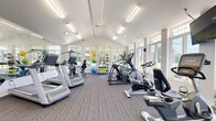 Located at 4905 W College Ave in Greendale, Wisconsin, this fitness center offers a welcoming and functional workout environment within a single, open-plan space. The room's most striking architectural feature is its vaulted ceiling, which lends a sense of height and openness well beyond what the footprint alone might suggest. Rows of large windows line the walls, flooding the interior with abundant natural light and providing pleasant views of the surrounding greenery outdoors.

The flooring throughout is durable, neutral-toned carpet tile, providing comfort underfoot during workouts. Overhead, flush fluorescent light fixtures supplement the natural light, ensuring consistent, bright illumination throughout the day. A full-length wall mirror on one side visually expands the space and allows users to monitor their form during exercise.

The fitness center is comprehensively equipped with a range of cardio machines, including Matrix treadmills, elliptical trainers, stationary and recumbent bikes, and additional aerobic equipment. A dedicated free weights area features a dumbbell rack, exercise balls in multiple sizes, and a cable/functional training station. A wall-mounted television is positioned for convenient viewing during cardio sessions.

Near the entrance, a windowed reception or office area, a coat rack, and framed informational displays complete the practical amenities of this well-appointed community fitness facility.