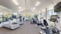 Located at 4905 W College Ave in Greendale, Wisconsin, this fitness center offers a welcoming and functional workout environment within a single, open-plan space. The room's most striking architectural feature is its vaulted ceiling, which lends a sense of height and openness well beyond what the footprint alone might suggest. Rows of large windows line the walls, flooding the interior with abundant natural light and providing pleasant views of the surrounding greenery outdoors.

The flooring throughout is durable, neutral-toned carpet tile, providing comfort underfoot during workouts. Overhead, flush fluorescent light fixtures supplement the natural light, ensuring consistent, bright illumination throughout the day. A full-length wall mirror on one side visually expands the space and allows users to monitor their form during exercise.

The fitness center is comprehensively equipped with a range of cardio machines, including Matrix treadmills, elliptical trainers, stationary and recumbent bikes, and additional aerobic equipment. A dedicated free weights area features a dumbbell rack, exercise balls in multiple sizes, and a cable/functional training station. A wall-mounted television is positioned for convenient viewing during cardio sessions.

Near the entrance, a windowed reception or office area, a coat rack, and framed informational displays complete the practical amenities of this well-appointed community fitness facility.