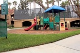 This outdoor playground offers a well-equipped recreational space designed for children's active play. The centerpiece is a multi-level play structure finished in bold green, red, and yellow, topped with a distinctive blue canopy roof that provides shade and a sense of enclosure. The structure incorporates a variety of engaging features, including a smooth red slide, a tunnel crawl-through element, climbing panels, and interactive activity boards, offering children multiple ways to explore and play.

The entire play area is surfaced with a generous layer of wood chip mulch, which serves as a cushioning safety base around all equipment. The playground is neatly bordered by a curved white edging that cleanly separates it from the surrounding well-manicured green lawn. Lush grass frames the perimeter, contributing to an open and inviting atmosphere.

Tall palm trees are visible throughout the surrounding landscape, lending a warm, sunny character to the setting. Residential and commercial buildings are visible in the background, indicating that this playground is situated within a community or hospitality complex. The overall environment is clean, well-maintained, and thoughtfully designed to provide a safe and engaging outdoor play experience.