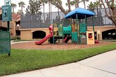 This outdoor playground offers a well-equipped recreational space designed for children's active play. The centerpiece is a multi-level play structure finished in bold green, red, and yellow, topped with a distinctive blue canopy roof that provides shade and a sense of enclosure. The structure incorporates a variety of engaging features, including a smooth red slide, a tunnel crawl-through element, climbing panels, and interactive activity boards, offering children multiple ways to explore and play.

The entire play area is surfaced with a generous layer of wood chip mulch, which serves as a cushioning safety base around all equipment. The playground is neatly bordered by a curved white edging that cleanly separates it from the surrounding well-manicured green lawn. Lush grass frames the perimeter, contributing to an open and inviting atmosphere.

Tall palm trees are visible throughout the surrounding landscape, lending a warm, sunny character to the setting. Residential and commercial buildings are visible in the background, indicating that this playground is situated within a community or hospitality complex. The overall environment is clean, well-maintained, and thoughtfully designed to provide a safe and engaging outdoor play experience.