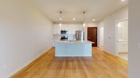 This well-appointed single-floor space is designed with a clean, modern aesthetic throughout. The heart of the home is an open-concept kitchen and living area, where warm hardwood flooring flows seamlessly underfoot. The kitchen is outfitted with crisp white cabinetry accented by brushed gold hardware, light stone countertops, a classic subway tile backsplash, and a full suite of stainless steel appliances including a refrigerator, microwave, and range. A central island with pendant lighting above serves as a natural gathering point and provides additional prep and seating space. The adjacent living area is bright and airy, benefiting from the open layout and a ceiling fan overhead. The bedroom features plush wall-to-wall carpeting, a ceiling fan, and a generously sized window that invites natural light and outdoor views. The bathroom is finished in neutral tones with large-format tile flooring, a white vanity with storage, and a tiled walk-in shower stall equipped with grab bars for added convenience. A dedicated laundry area houses a stacked front-loading washer and dryer alongside built-in wire shelving for added storage. Throughout the space, a cohesive palette of warm whites and soft neutrals creates a calm, functional living environment.