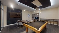 This contemporary amenity lounge is designed with both recreation and relaxation in mind, offering a well-appointed, single-level space with a cohesive modern aesthetic throughout. The centerpiece of the room is a full-size pool table with a sleek black felt surface and warm natural wood frame, positioned beneath a dramatic pendant light fixture that cascades multiple Edison-style bulbs from a bold black recessed ceiling detail — creating a striking focal point that anchors the entire space.

Dark, large-format tile flooring runs throughout, providing an elegant contrast against the neutral cream-toned walls. A dark accent wall fitted with a wall-mounted flat-screen television defines the lounge zone, which includes a round dark wood dining table surrounded by contemporary gray chairs, as well as a casual seating arrangement featuring cream upholstered chairs and a small marble-top side table.

Along one wall, a built-in wet bar features wood-grain cabinetry, a dark countertop, sink, and microwave — ideal for entertaining. A high-top bar counter with seating lines the adjacent wall, offering additional social space. Abstract framed artwork is thoughtfully placed throughout, reinforcing the room's refined, gallery-like atmosphere.

Floor-to-ceiling glass partitions with aluminum framing separate the lounge from a connecting corridor, maintaining visual openness while defining distinct zones within the space.