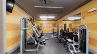 This fitness center is a compact yet fully functional workout space designed to accommodate a range of exercise needs. The room features warm, horizontally striped yellow and orange walls that lend an energetic ambiance, complemented by durable dark rubber flooring suited for high-traffic use. Overhead fluorescent lighting ensures the space is well-lit throughout, while several windows with wooden blinds allow for natural light to filter in.

The layout is efficiently organized, with cardio equipment such as ellipticals and steppers positioned along one side, and strength training machines arranged throughout the room. A free weights area includes a rack with multiple dumbbell options and a flat bench, catering to both beginner and experienced gym-goers. A wall-mounted television is available for entertainment during workouts.

Full-length mirrors line one wall, visually expanding the space and allowing users to monitor their form. A wall clock keeps exercisers on schedule. The overall design prioritizes practicality, making it a convenient and accessible fitness amenity.