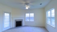 This single-floor residential space is thoughtfully designed for comfortable everyday living. The main living area serves as the heart of the home, showcasing a classic tile-surround fireplace with a crisp white mantel that provides a clean, traditional focal point to the room. Soft, neutral-toned carpeting covers the floor, adding warmth and a sense of comfort underfoot. A ceiling fan with an integrated light fixture is centrally positioned overhead, offering both practical air circulation and ambient lighting throughout the space. Large double-hung windows allow generous amounts of natural light to fill the room, illuminating the soft, light-colored walls and creating an airy, inviting atmosphere. The windows also frame views of the surrounding outdoor scenery, connecting the interior to the natural environment beyond. The overall aesthetic is clean and understated, with neutral tones throughout that provide a versatile backdrop suited to a variety of interior design styles. The layout is open and uncluttered, allowing for flexible furniture arrangements to suit a range of living needs. A ceiling vent indicates a central HVAC system, ensuring year-round climate control. This space offers a welcoming and functional environment well-suited for relaxed, daily living.