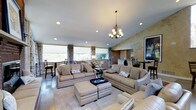 This single-floor space showcases a thoughtfully designed open-concept layout that seamlessly connects the living, dining, and kitchen areas. The living room is a standout centerpiece, featuring vaulted ceilings accented by a decorative chandelier and complemented by recessed lighting throughout. A textured accent wall in warm earth tones adds depth and character to the space, while a framed artwork piece anchors the room with a refined aesthetic.

The living area is generously proportioned, accommodating a full suite of plush, neutral-toned sofas and ottomans arranged around a central area rug, creating an inviting and comfortable gathering space. Large windows dressed with layered curtains and roller shades line the walls, flooding the interior with natural light and offering views of the surrounding outdoor landscape.

Adjacent to the living room, a dining area with a bistro-style table and chairs provides an intimate setting for meals. Further back, a glimpse of the kitchen reveals a light-colored counter and cabinetry, suggesting a clean and functional culinary space. The polished hard flooring throughout ties the areas together cohesively, enhancing the open and airy ambiance that defines this well-appointed single-story space.