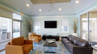 This business center lounge is designed to offer a welcoming and functional environment for work and relaxation. The space is distinguished by its tray ceiling with recessed lighting and a ceiling fan, adding both architectural interest and practical comfort. Soft mint-green walls create a calm, professional atmosphere throughout the room.

The seating area is thoughtfully arranged, featuring a dark leather sofa paired with warm cognac leather armchairs, all centered around a sleek glass-and-wood coffee table. A patterned area rug in shades of blue and green anchors the furniture grouping, adding texture and visual depth to the space.

A wall-mounted flat-screen television is positioned centrally above a dark wood media console, making it a natural focal point. Flanking the TV, two windows dressed with white roller shades allow natural light to filter in while maintaining privacy. Decorative dried grasses in tall vases add organic touches to the corners of the room, enhancing the overall aesthetic.

The single-floor layout ensures easy accessibility, and the cohesive design balances professional functionality with a comfortable, inviting ambiance suitable for both individual work and informal gatherings.