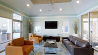 This business center lounge is designed to offer a welcoming and functional environment for work and relaxation. The space is distinguished by its tray ceiling with recessed lighting and a ceiling fan, adding both architectural interest and practical comfort. Soft mint-green walls create a calm, professional atmosphere throughout the room.

The seating area is thoughtfully arranged, featuring a dark leather sofa paired with warm cognac leather armchairs, all centered around a sleek glass-and-wood coffee table. A patterned area rug in shades of blue and green anchors the furniture grouping, adding texture and visual depth to the space.

A wall-mounted flat-screen television is positioned centrally above a dark wood media console, making it a natural focal point. Flanking the TV, two windows dressed with white roller shades allow natural light to filter in while maintaining privacy. Decorative dried grasses in tall vases add organic touches to the corners of the room, enhancing the overall aesthetic.

The single-floor layout ensures easy accessibility, and the cohesive design balances professional functionality with a comfortable, inviting ambiance suitable for both individual work and informal gatherings.